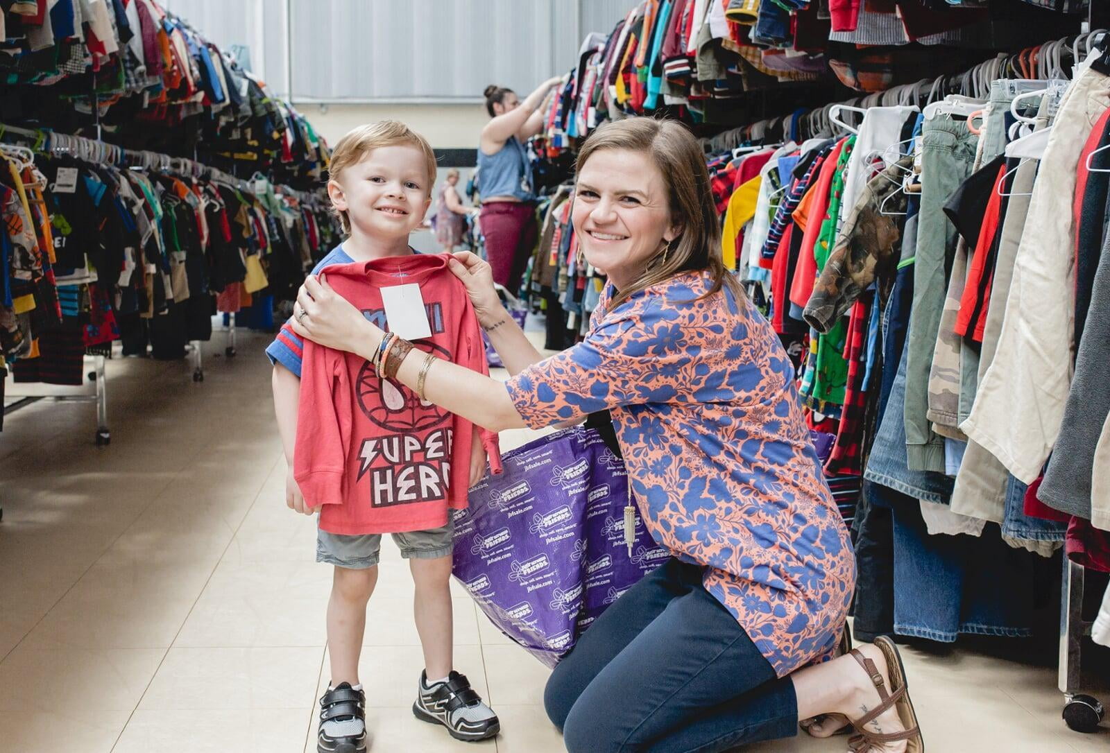A mom with a large JBF shopping bag on her shoulder stands beside her husband who wears their toddler at a JBF sale.